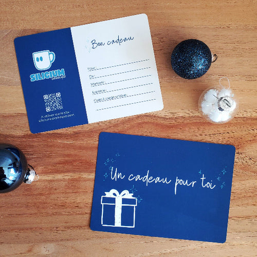 Carte cadeau de la taille d'une carte postale avec un côté sur lequel est inscrit "Un cadeau pour toi" et de l'autre côté, les informations "Pour", "De", "Montant", "Date d'expiration" et "Code cadeau" sont disponibles pour être complétés. Un QR permettant d'accéder au site web est également présent sur cette face.