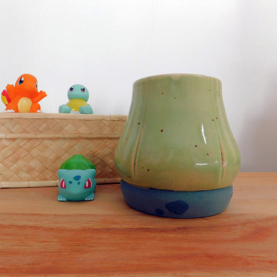 Tasse en grès blanc faisant référence à Bulbizarre. La tasse reprend la forme du bulbe sur le dos du Pokémon et est émaillé en vert clair. Le bulbe repose sur un base vert bleu pâle avec des tâches vert émeraude comme le corps de Bulbizarre. La anse est fait référence aux deux lianes du Pokémon.