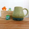 Tasse en grès blanc faisant référence à Bulbizarre. La tasse reprend la forme du bulbe sur le dos du Pokémon et est émaillé en vert clair. Le bulbe repose sur un base vert bleu pâle avec des tâches vert émeraude comme le corps de Bulbizarre. La anse est fait référence aux deux lianes du Pokémon.
