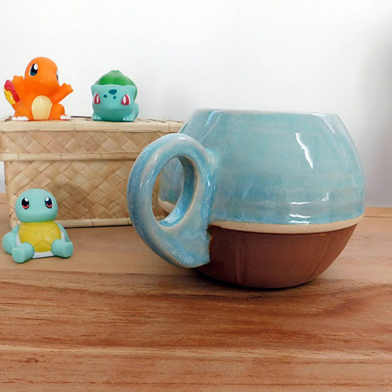 Tasse faisant référence à Carapuce. La tasse est arrondie, le bas représente une carapace de tortue marron foncé et le haut est émaillé en bleu clair avec un effet eau.