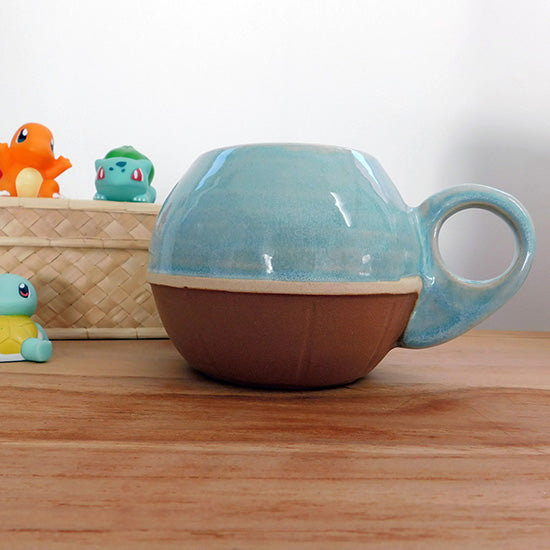 Tasse en grès blanc faisant référence à Carapuce. La tasse est arrondie, le bas représente une carapace de tortue marron foncé et le haut est émaillé en bleu clair avec un effet eau. La anse de la tasse rappelle la queue de Carapuce et est percée pour permettre le passage d'un ou deux doigts.