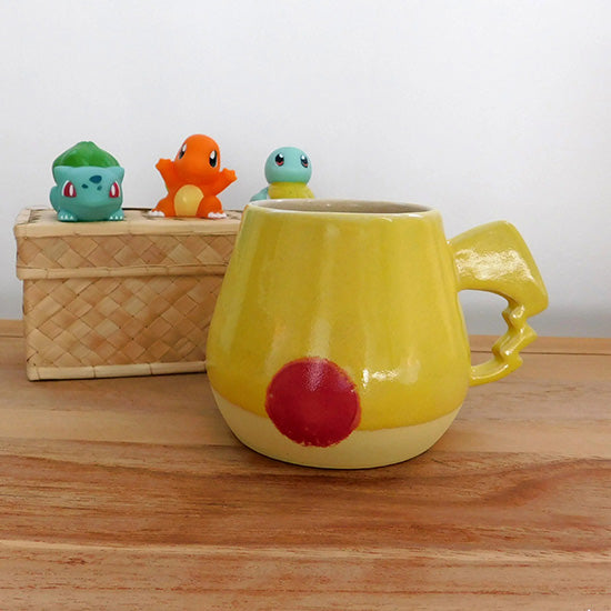 Tasse en grès blanc faisant référence à Pikachu. La tasse est reprend la forme du visage de Pikachu, elle colorée en jaune matte à la base et en jaune brillant sur la partie supérieur.  On retrouve les joues rouges de Pikachu et la anse est en forme d'éclair.