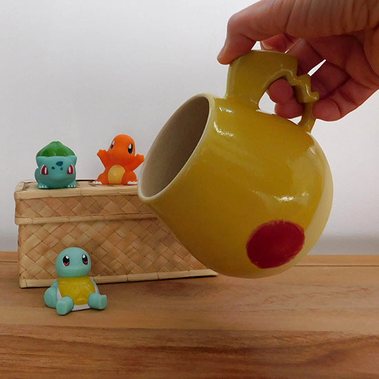 Tasse en grès blanc faisant référence à Pikachu. La tasse est reprend la forme du visage de Pikachu, elle colorée en jaune matte à la base et en jaune brillant sur la partie supérieur.  On retrouve les joues rouges de Pikachu et la anse est en forme d'éclair.