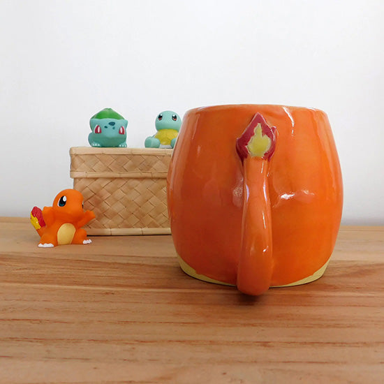Tasse en grès blanc faisant référence à Salamèche. La tasse est en forme  d'olive et émaillée en orange sur le haut. Elle est colorée en jaune matte sur le bas pour rappeler le ventre du pokémon. La anse reprend la forme de la queue de Salamèche avec sa flamme.